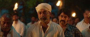 Movie still from “Kantara” (2022), directed by Rishab Shetty – A man with a white turban is standing in front of a group of people; Close Up shot, Over the shoulder angle