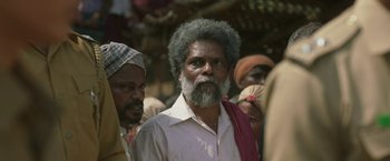 Movie still from “Kantara” (2022), directed by Rishab Shetty – A man with a beard and a white shirt is standing in a crowd; Close Up shot, Over the shoulder angle