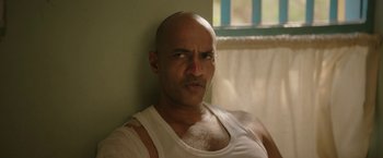 Movie still from “Kantara” (2022), directed by Rishab Shetty – A bald man in a white tank top looking at the camera; Close Up shot, Over the shoulder angle