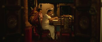 Movie still from “Kantara” (2022), directed by Rishab Shetty – A man sitting at a dinner table in front of a woman; Wide shot, High angle
