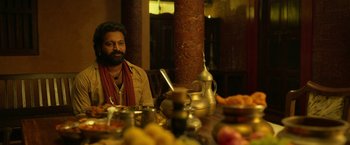 Movie still from “Kantara” (2022), directed by Rishab Shetty – A man sitting at a table with a bowl of food; Medium shot, Over the shoulder angle