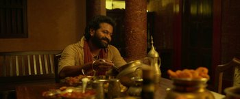 Movie still from “Kantara” (2022), directed by Rishab Shetty – A man sitting at a wooden table with a pot; Close Up shot, Over the shoulder angle