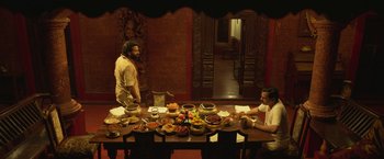 Movie still from “Kantara” (2022), directed by Rishab Shetty – Two men sitting at a long table with food on top of it; Wide shot, High angle