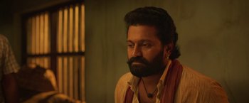 Movie still from “Kantara” (2022), directed by Rishab Shetty – A man with a beard is sitting in a dimly lit room; Close Up shot, Low angle