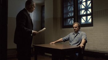 Movie still from “Kill the Irishman” (2011), directed by Jonathan Hensleigh – A man sitting at a table in front of another man holding papers; Medium shot, Over the shoulder angle
