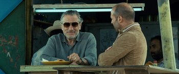 Movie still from “Killer Elite” (2011), directed by Gary McKendry – A man sitting at a table talking to another man; Close Up shot, Over the shoulder angle