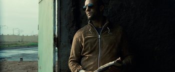 Movie still from “Killer Elite” (2011), directed by Gary McKendry – A man in a brown jacket and sunglasses holding a book; Close Up shot, Low angle