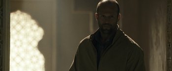 Movie still from “Killer Elite” (2011), directed by Gary McKendry – A bald man with a beard is standing in a room; Close Up shot, Low angle