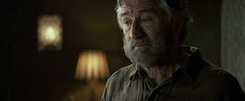 Movie still from “Killer Elite” (2011), directed by Gary McKendry – An older man with a beard and a gray shirt; Close Up shot, Over the shoulder angle