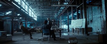 Movie still from “Killer Elite” (2011), directed by Gary McKendry – Two people sitting at a table in a warehouse; Wide shot, Over the shoulder angle