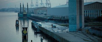 Movie still from “Killer Elite” (2011), directed by Gary McKendry – A man standing next to a boat on a body of water; Extreme Wide shot, High angle