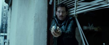 Movie still from “Killer Elite” (2011), directed by Gary McKendry – A man in a leather jacket holding a gun in his hand; Medium shot, Low angle