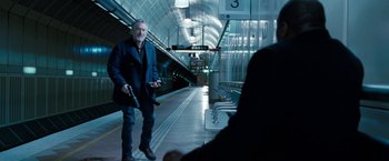 Movie still from “Killer Elite” (2011), directed by Gary McKendry – A man holding a gun while standing on a train platform; Wide shot, Over the shoulder angle