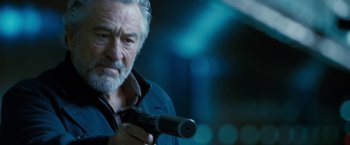 Movie still from “Killer Elite” (2011), directed by Gary McKendry – A man holding a gun while standing in a room; Close Up shot, Over the shoulder angle