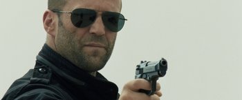 Movie still from “Killer Elite” (2011), directed by Gary McKendry – A person holding a gun wearing sunglasses; Close Up shot, Low angle
