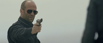 Movie still from “Killer Elite” (2011), directed by Gary McKendry – A man in a black jacket and sunglasses holding a gun; Close Up shot, Low angle
