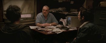 Movie still from “Killing Them Softly” (2012), directed by Andrew Dominik – A man sitting at a table with papers on the table; Medium shot, Over the shoulder angle
