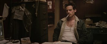 Movie still from “Killing Them Softly” (2012), directed by Andrew Dominik – A man sitting at a desk smoking a cigarette in front of an open filing cabinet; Medium shot, Over the shoulder angle