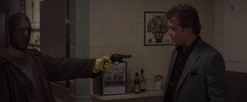 Movie still from “Killing Them Softly” (2012), directed by Andrew Dominik – A man holding a gun pointed at another man in a kitchen; Medium shot, Over the shoulder angle