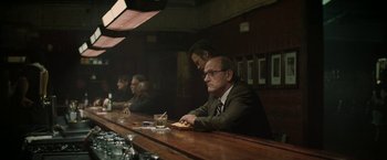 Movie still from “Killing Them Softly” (2012), directed by Andrew Dominik – A man sitting at a bar with other people; Medium shot, Over the shoulder angle