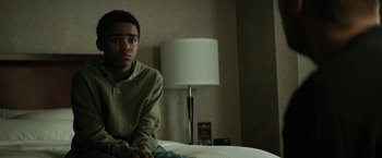 Movie still from “Kin” (2018), directed by Josh Baker – A young man sitting on a bed in front of a lamp; Medium shot, Low angle