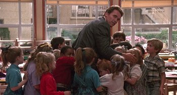 Movie still from “Kindergarten Cop” (1990), directed by Ivan Reitman – A group of children are gathered around a man in a suit; Medium shot, Over the shoulder angle