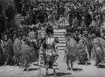 Movie still from “King Kong” (1933), directed by Merian C. Cooper – A group of people in costumes and masks in front of a crowd of onlookers; Wide shot, High angle