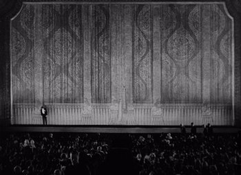 Movie still from “King Kong” (1933), directed by Merian C. Cooper – A man standing on a stage in front of an audience; Extreme Wide shot, High angle