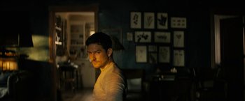 Movie still from “Knock at the Cabin” (2023), directed by M. Night Shyamalan – A man standing in a living room in front of pictures; Medium shot, Low angle