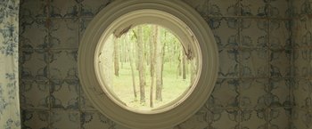 Movie still from “Knock at the Cabin” (2023), directed by M. Night Shyamalan – A round window in a room with a forest in the background; Extreme Wide shot, Overhead angle