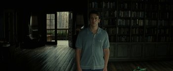 Movie still from “Knock at the Cabin” (2023), directed by M. Night Shyamalan – A man standing in front of a bookcase in a room; Medium shot, Low angle