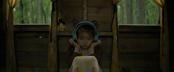 Movie still from “Knock at the Cabin” (2023), directed by M. Night Shyamalan – A little girl wearing headphones while sitting in a chair; Medium shot, High angle