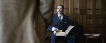 Movie still from “Kon-Tiki” (2012), directed by Espen Sandberg – A man in a suit sitting in a chair holding a box; Medium shot, Over the shoulder angle