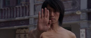 A shirtless man with long black hair holding up his hand