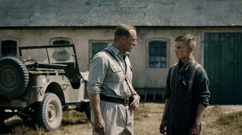 Movie still from “Land of Mine” (2015), directed by Martin Zandvliet – A man and a boy standing next to each other in front of a building; Medium shot, Over the shoulder angle