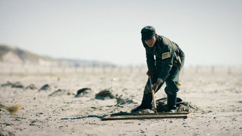 Movie still from “Land of Mine” (2015), directed by Martin Zandvliet – A man in a hat is digging a hole in the sand; Wide shot, High angle