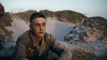 Movie still from “Land of Mine” (2015), directed by Martin Zandvliet – A man sitting in the grass in front of some sand dunes; Close Up shot, Over the shoulder angle