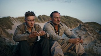 Movie still from “Land of Mine” (2015), directed by Martin Zandvliet – A couple of men sitting next to each other on a beach; Medium shot, Over the shoulder angle