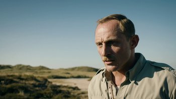 Movie still from “Land of Mine” (2015), directed by Martin Zandvliet – A person wearing a shirt near some sand dunes; Close Up shot, Over the shoulder angle