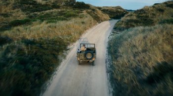 Movie still from “Land of Mine” (2015), directed by Martin Zandvliet – A man riding in the back of a jeep down a dirt road; Extreme Wide shot, High angle