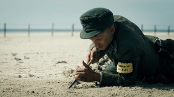 Movie still from “Land of Mine” (2015), directed by Martin Zandvliet – A man in a military uniform on the beach writing; Medium shot, High angle
