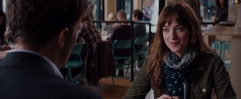 Movie still from “Fifty Shades of Grey” (2015), directed by Sam Taylor-Johnson – A woman sitting at a table in front of other people; Medium shot, Over the shoulder angle
