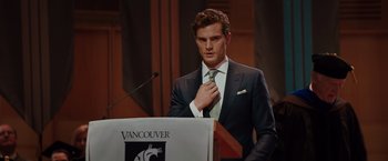 Movie still from “Fifty Shades of Grey” (2015), directed by Sam Taylor-Johnson – A man adjusts his neck tie at a podium; Medium shot, Over the shoulder angle