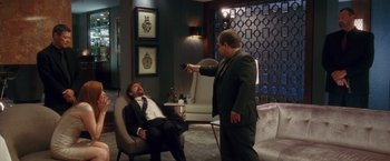 Movie still from “Keeping Up with the Joneses” (2016), directed by Greg Mottola – A man in a suit and tie holding a remote control; Wide shot, Over the shoulder angle