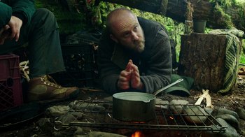 Movie still from “Leave No Trace” (2018), directed by Debra Granik – A man with a beard is sitting on the ground near a pot of food; Medium shot, Low angle
