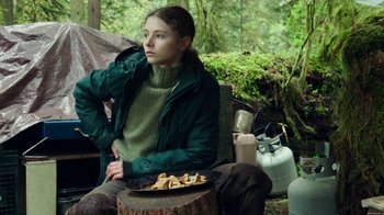 Movie still from “Leave No Trace” (2018), directed by Debra Granik – A woman sitting on a tree stump in the woods; Medium shot, Over the shoulder angle