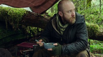 Movie still from “Leave No Trace” (2018), directed by Debra Granik – A man sitting in the woods holding a bowl of food; Medium shot, Low angle