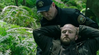 Movie still from “Leave No Trace” (2018), directed by Debra Granik – A man in a black jacket is being held by a police officer; Medium shot, Low angle