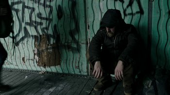 Movie still from “Leave No Trace” (2018), directed by Debra Granik – A man squatting on the ground in front of a wall; Medium shot, High angle