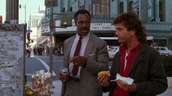 Movie still from “Lethal Weapon” (1987), directed by Richard Donner – Two men standing on the side of the street eating food; Medium shot, Over the shoulder angle